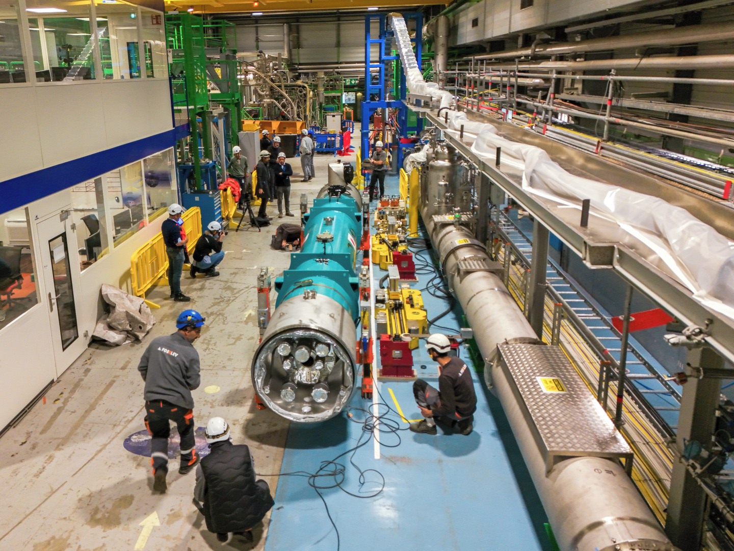 View of an industrial hall with many persons with helmets and a bige blue tube (the magnet) in the middle.